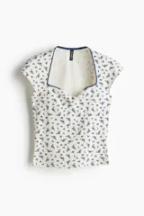 Top corto de punto acanalado fino de algodón color crema con estampado floral azul, mangas cortas y escote corazón con ribete de picot azul.