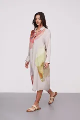 Vestido camisero midi color beige con estampado abstracto en tonos naranja, rojo, verde y amarillo. Tiene cuello camisero, mangas largas con puños abotonados y cierre frontal con botones.