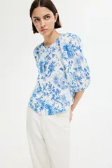 Blusa blanca con estampado floral azul, cuello redondo, cierre de botones y mangas globo tres cuartos con puños elásticos.