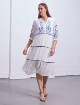 Vestido midi blanco con bordados florales azules en el escote, mangas y cintura. Tiene cuello en V, mangas 3/4 con botón y falda escalonada con detalles de ribetes azules.