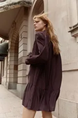 Vestido corto color marrón oscuro, estilo camisero, con cuello mao con ribete de volado y tapeta de botones con el mismo detalle. Posee mangas largas con puños sencillos de volado y un cinturón fino extraíble para ajustar la cintura. La falda tiene un fruncido escalonado que le da vuelo.