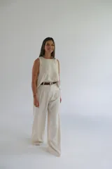 Pantalón de vestir color crudo, de corte palazzo y tiro alto. Presenta pinzas en la cintura y cierre frontal con botón y cremallera. Incluye cinturón trenzado marrón con hebilla redonda plateada.