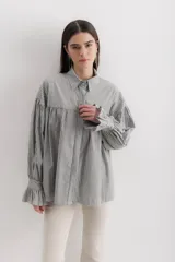 Camisa gris a rayas verticales blancas, de corte amplio, cuello alto con lazo y mangas largas abullonadas con puños elásticos.