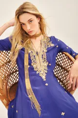 Vestido largo azul vibrante con escote en V profundo, mangas 3/4 y caída fluida. Presenta un elaborado bordado frontal en el escote con piedras, cintas e hilos de lurex en tonos dorados y plateados, con pequeños apliques florales dorados distribuidos en el cuerpo de la prenda. Incluye un accesorio colgante de cuentas doradas a modo de bufanda o detalle decorativo.