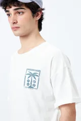 Remera color beige de corte amplio con mangas cortas y estampado de una postal con una palmera en el pecho.