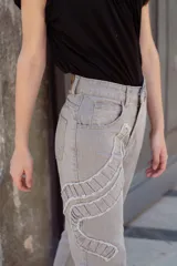 Jean gris lavado, estilo mom fit, con aplique fruncido en forma de espiral en la pierna izquierda, tiro alto, bolsillos delanteros y traseros funcionales, y cierre frontal con botón y cremallera.