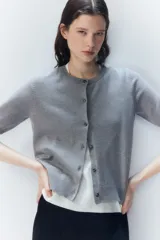 Cárdigan gris de punto fino con cuello redondo, manga corta y cierre frontal con botones. Tiene terminaciones acanaladas en cuello, puños y ruedo.