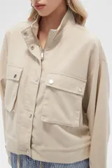 Campera color beige de manga larga, con cuello alto, cierre frontal con cremallera y botones a presión, bolsillos con solapa en el pecho y puños con trabilla ajustable.