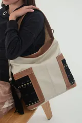 Bolso tipo tote color crudo con detalles en cuero color terracota y apliques de gamuza negra bordada con tachas. Tiene correa de hombro y cierre metálico.