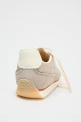 Zapatillas deportivas tipo running, color gris claro con detalles en beige y blanco. Combinación de materiales en el corte y suela en contraste. Cierre mediante cordones de color.