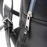 Mochila negra de cuero sintético con cierre superior y bolsillo frontal con cierre. Tiene asa de mano fija, correas regulables, herrajes plateados y logo de la marca en el frente. El interior está forrado en tela de algodón y poliéster y cuenta con un bolsillo con cierre y dos bolsillos abiertos.