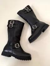 Bota de caña media de cuero vacuno color marrón oscuro, con punta cuadrada, cierre lateral y suela track negra. Presenta detalles de hebillas metálicas plateadas en la caña y en la parte inferior del empeine.