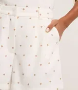 Bermuda de piquet color blanco con estampado de lunares negros, de corte recto, tiro alto, con trabillas en la cintura, pliegues y cierre tradicional.