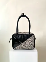 Cartera rígida artesanal de cuero con tachas plateadas, herrajes y cierre metálico plateado YKK. Tiene asa fina para llevar al hombro o en la mano y correa larga extra regulable. El interior es forrado, tiene un bolsillo con cierre enmarcado en cuero, y un segundo bolsillo plaqué.