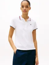 Chomba blanca de corte slim fit, confeccionada en algodón elástico. Presenta cuello y mangas con ribetes en azul y rojo, tapeta de cinco botones y logo de Tommy Hilfiger bordado en el pecho.