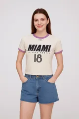 Remera corta color crema con cuello redondo y mangas cortas con ribete lila. Estampado frontal con la palabra "Miami" en negro, la leyenda "Since 2001" y el número 18.