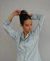 Camisa celeste de satén con cuello clásico, manga larga con puños abotonados y cierre frontal con botones. Tiene un bolsillo de parche en el pecho y detalles de ribete en contraste.