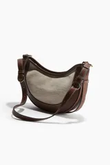 Cartera marrón con panel frontal de lona color crema, forma de media luna y correa ajustable.