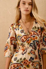 Vestido corto estampado con motivos florales en tonos naranja, azul y verde sobre fondo blanco. Tiene mangas 3/4 con volados en los puños, escote en V y cintura elastizada con lazo.