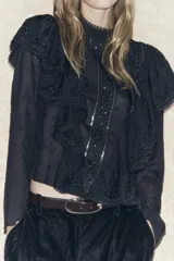 Blusa negra de viscosa con cuello perkins y manga larga. Detalles de bordados, lentejuelas y volantes. Cierre en la espalda con abertura y botones.