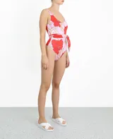 Traje de baño enterizo con estampado de flores rojas y hojas color crema sobre fondo lila. Presenta corte alto en la pierna y cinturón lila con lazada lateral.