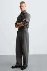 Pantalón negro de corte amplio con cinturón de tela con hebilla metálica. Presenta pliegues frontales, bolsillos laterales y traseros. Cierre frontal con cremallera y botón.