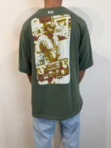 Remera verde militar de corte oversize con estampado rectangular en la espalda con imagen de persona con equipo de música.