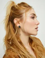 Pendientes colgantes bañados en oro 18k. Cada pendiente presenta un cristal facetado ovalado de color gris oscuro engastado, del cual cuelgan dos eslabones alargados de metal dorado.