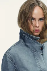 Chaqueta de denim celeste con cuello alto, manga larga con trabilla y bolsillos laterales. Presenta detalle de pliegues en la espalda y cierre frontal con botones ocultos.