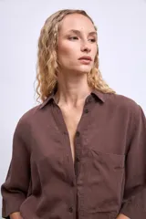 Camisa blanca de manga tres cuartos, con cuello camisero y corte holgado. Presenta un bolsillo de parche en el pecho izquierdo.