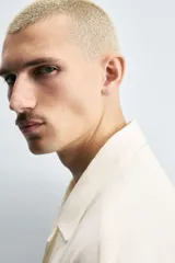 Camisa de denim de algodón, color beige claro, con corte relajado, cuello bowling y manga corta. Presenta cierre frontal con botones a presión y un pequeño bordado a tono en el pecho.