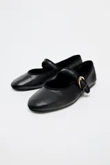 Zapato plano tipo bailarina color negro, confeccionado en piel, con tira delantera con hebilla metálica ajustable y acabado en punta redonda.
