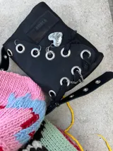 Cartera de cuero negra con correa regulable, estilo bandolera o al hombro. Decorada con charm de corazón metálico, encaje negro, cadenas y aros metálicos.