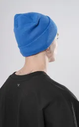 Gorro de tejido liso color azul eléctrico, con borde doblado y un pequeño parche cuadrado negro con un logo bordado en blanco que simula unas tijeras cruzadas.