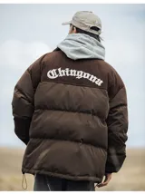 Campera puffer marrón con paneles horizontales acolchados. La parte superior de la espalda es de pana marrón con el texto 'Chingona' bordado en blanco. Posee cordón ajustable en el ruedo y se lleva superpuesta a una sudadera con capucha gris.