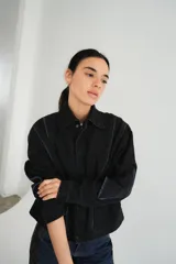 Campera de jean negra tipo bomber con cuello redondo y cierre frontal con botones metálicos. Presenta un parche rectangular con diseño gráfico en la espalda.