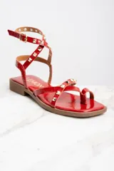 Sandalias rojas de charol con tiras finas, puntera cuadrada y cierre con hebilla al tobillo. Decoradas con tachas piramidales doradas.