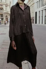 Camisa oversize color marrón oscuro confeccionada en tejido satinado con efecto arrugado, cuello solapa, manga larga con puños y cierre frontal con botones metálicos.