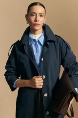 Trench color beige de corte clásico con cuello amplio, botones frontales, detalles en los hombros y mangas largas.