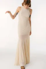 Vestido largo color beige, con cuello alto y sin mangas. La parte superior es ajustada al cuerpo y la falda es amplia y fluida, con paneles traslúcidos.