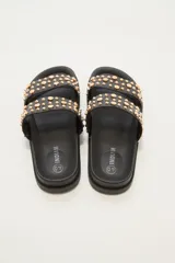 Sandalia tipo slide de color crudo, con dos tiras anchas adornadas con tachas doradas redondas de diferentes tamaños. La suela es gruesa y de goma.