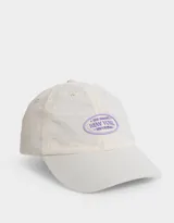 Gorra de gabardina color crudo con visera rígida y aplique ovalado de silicona color lila con texto.