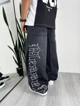 Pantalón de jean negro lavado, corte muy ancho (baggy) con estampado vertical en blanco con tipografía gótica en el lateral de la pierna.
