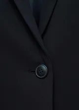 Chaqueta negra con cuello de pico con solapa y cierre de botón en la parte delantera. Tiene manga larga con puños abotonados, diseño recto, estilo traje y dos bolsillos de solapa en la parte delantera.
