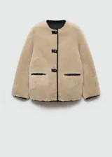 Chaqueta beige de poliéster con textura de borreguito, cuello redondo, cierre frontal con botones tipo trenca, ribetes en contraste de color negro, dos bolsillos delanteros y forro interior.