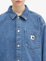 Camisa de denim azul de Carhartt WIP, de corte holgado, manga larga, cuello clásico y cierre frontal con botones. Tiene un bolsillo en el pecho con etiqueta con el logo de la marca.