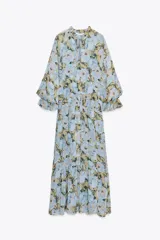 Vestido largo con estampado floral en tonos celeste, amarillo y marrón. Tiene cuello redondo con volantes y lazada, escote pico, manga larga con puño elástico con volantes, forro interior y cierre frontal con botones.