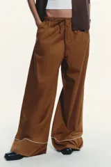 Pantalón marrón de popelina con corte palazzo, tiro medio, cintura elástica ajustable con cordones, bolsillos laterales y falsos bolsillos de vivo con botones en la espalda. Detalle de vivos blancos a contraste en el ruedo.