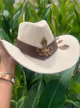 Sombrero de ala ancha tejido en hilo de coco y sintético, color marrón claro. La copa está cubierta con encaje naranja quemado con patrón floral. Tiene una cinta de cuero marrón oscuro y un aplique decorativo de hoja de gamuza con incrustaciones de pedrería.