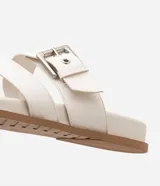 Sandalias planas de cuero sintético color blanco, con puntera redonda y dos tiras, una con hebilla metálica y otra en forma de H.
